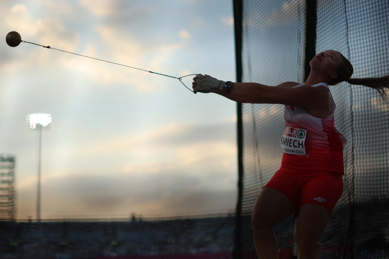 Aleksandra Śmiech w finale Lekkoatletka w czerwono-białym stroju wykonuje rzut młotem na stadionie sportowym podczas zawodów, trzymając sprzęt obiema rękami w dynamicznej pozie na tle wieczornego nieba.