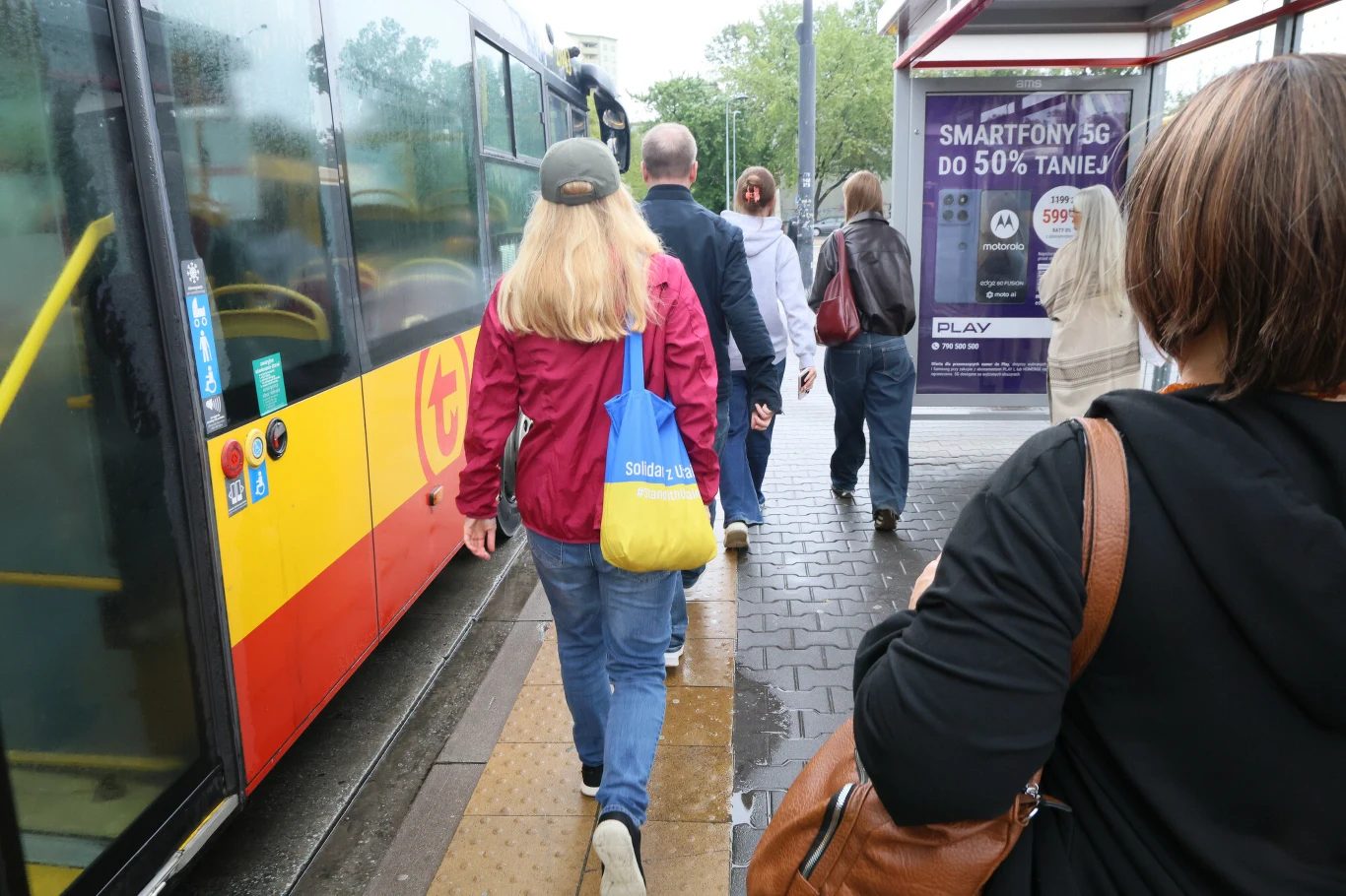 Coraz więcej Ukraińców opuszcza Polskę Grupa osób wchodzi do miejskiego autobusu, widoczny przystanek z wiatą i reklamą operatora komórkowego.