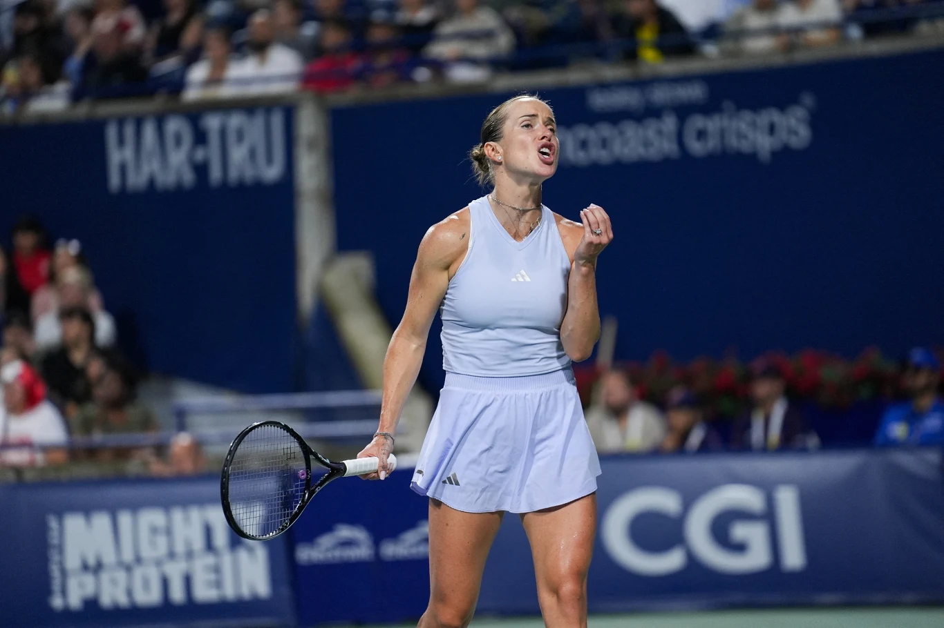 Elina Switolina Tenisistka w jasnej sportowej sukience z zaciśniętą pięścią i wyrazem determinacji na twarzy w trakcie meczu na korcie, w tle widoczna rozmyta publiczność i reklamy.
