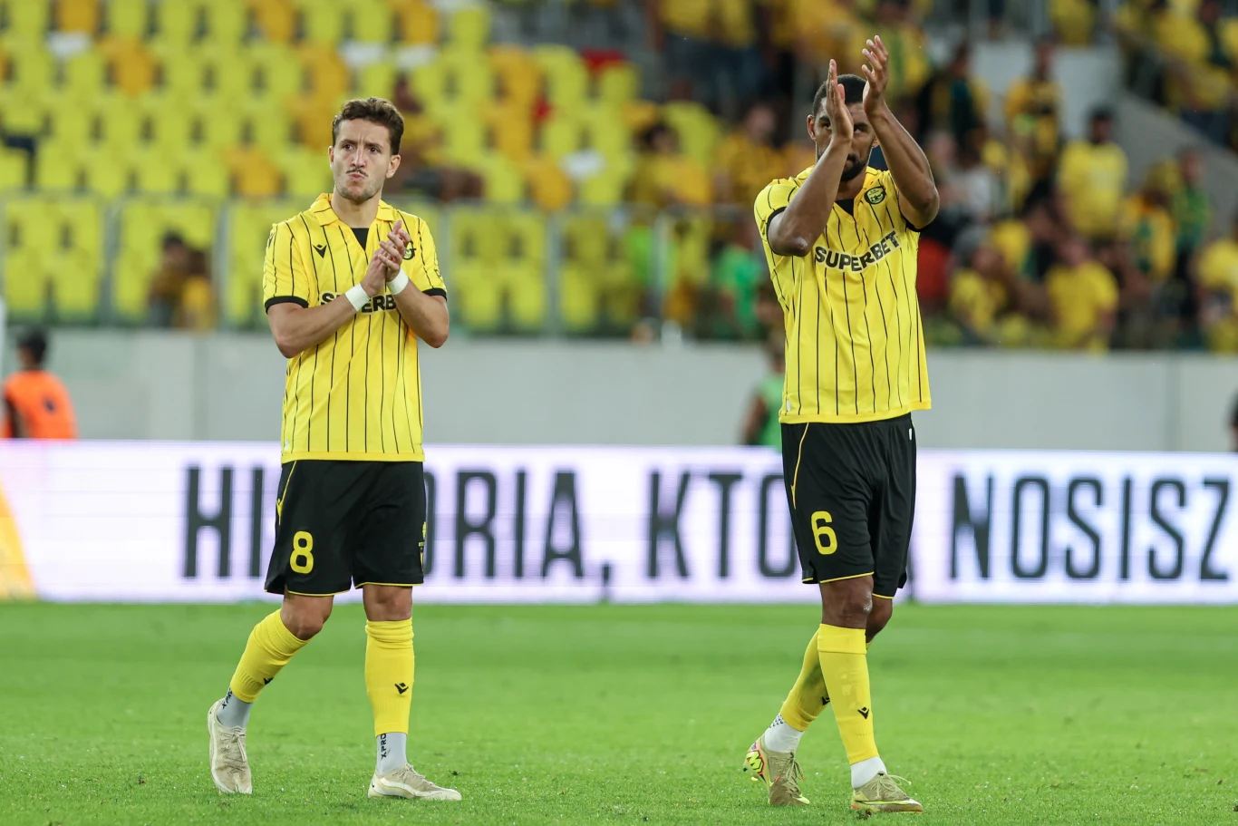 Borja Galan i Lukas Klemenz w barwach GKS-u Katowice Dwóch piłkarzy w żółto-czarnych strojach z numerami 8 i 6 klaszcze, stojąc na murawie stadionu podczas meczu, w tle trybuny wypełnione kibicami.