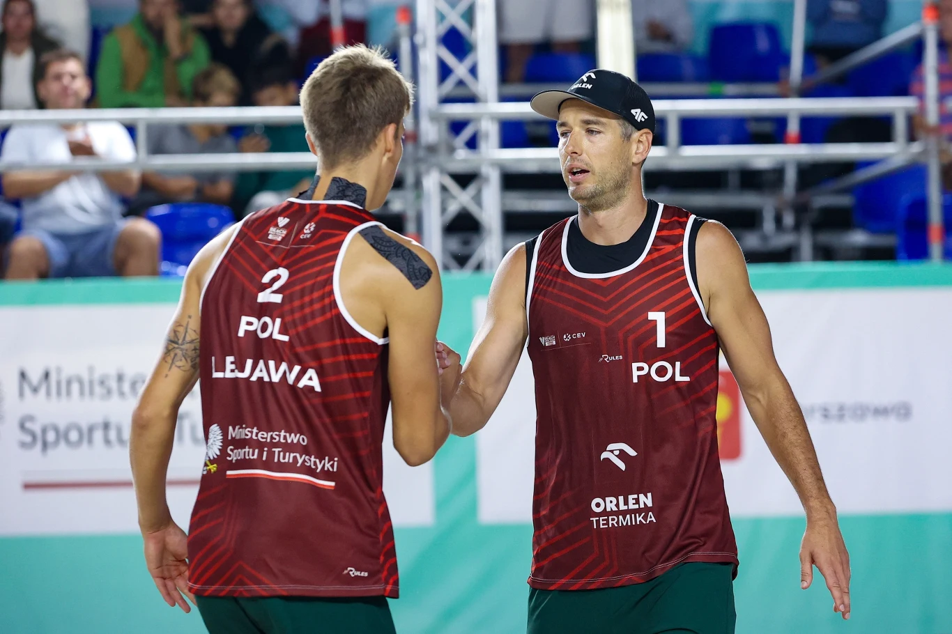Piotr Kantor (z prawej) i Filip Lejawa Piotr Kantor i Filip Lejawa na boisku do siatkówki plażowej, wymieniają uścisk dłoni, czerwone koszulki POL.