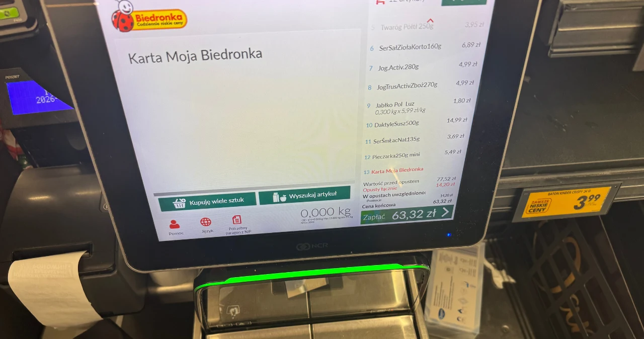 Biedronka odpala ulubione promocje Polaków. Duże obniżki na mięso, masło i kawę Ekran kasy samoobsługowej Biedronki z listą produktów i kwotą 63,32 zł, widoczny terminal płatniczy i paragon.