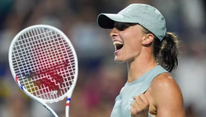 Tenisistka w sportowej czapce i szarym stroju unosi zwycięsko zaciśniętą pięść, trzymając rakietę tenisową, z szerokim uśmiechem wyrażającym radość po wygranym punkcie.