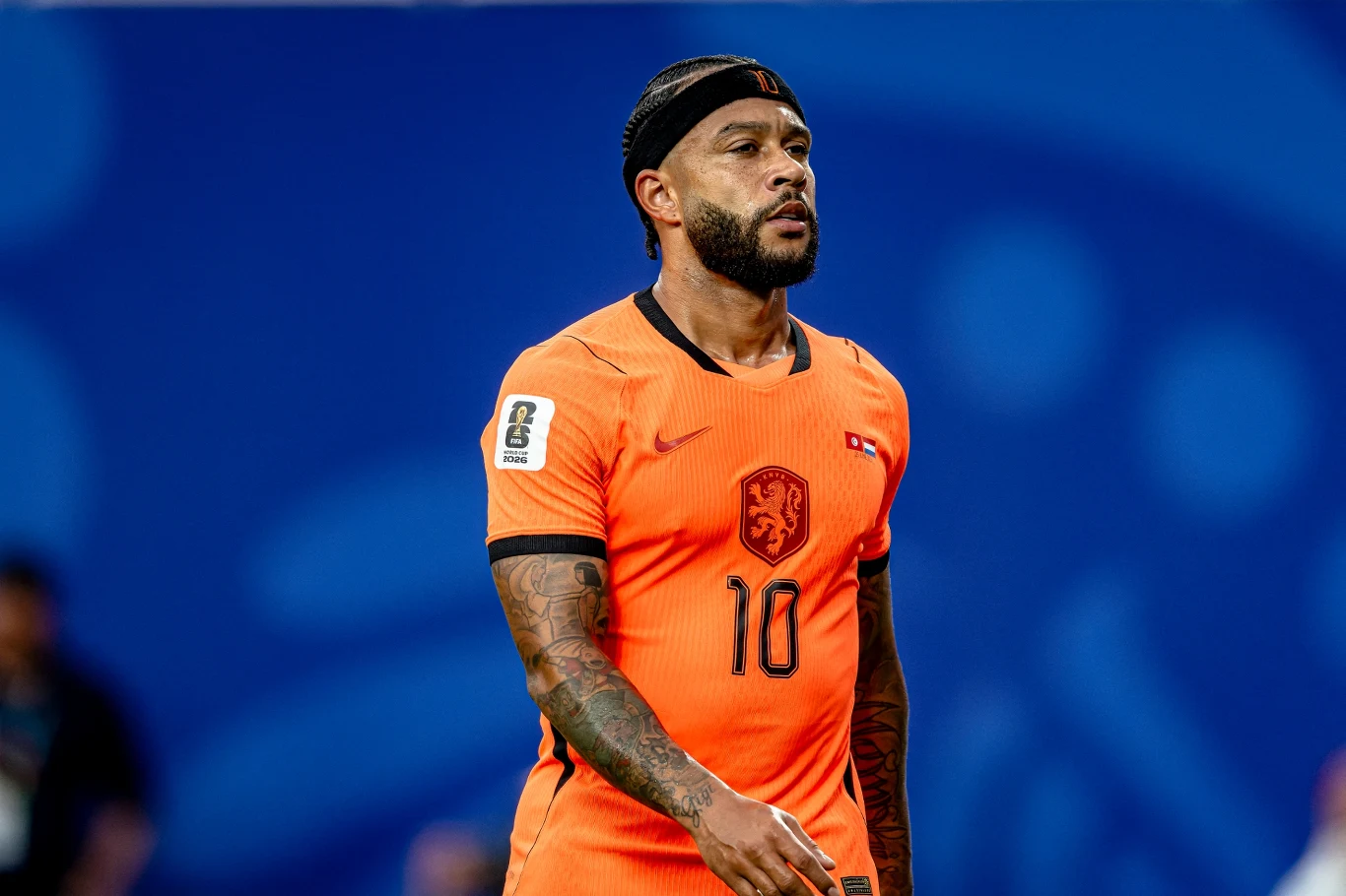 Memphis Depay Piłkarz w pomarańczowej koszulce z numerem 10 i opaską na głowie podczas meczu piłkarskiego na niebieskim tle stadionu.