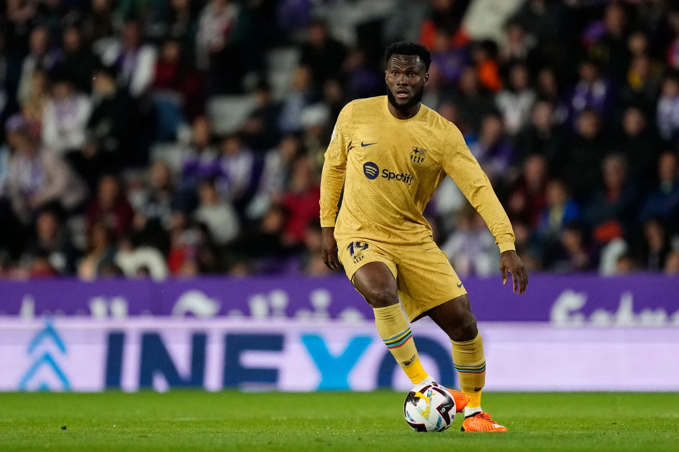 Franck Kessie w barwach Barcelony Piłkarz w złotym stroju FC Barcelony prowadzi piłkę po murawie podczas meczu, skoncentrowany na grze, w tle zamazana trybuna z kibicami.