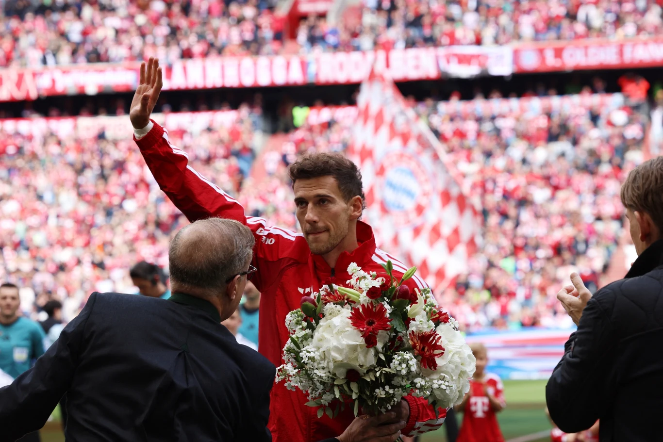 Leon Goretzka Piłkarz w czerwonym stroju trzymający bukiet kwiatów i unoszący rękę w geście pozdrowienia, otoczony przez tłum ludzi na trybunach stadionu.