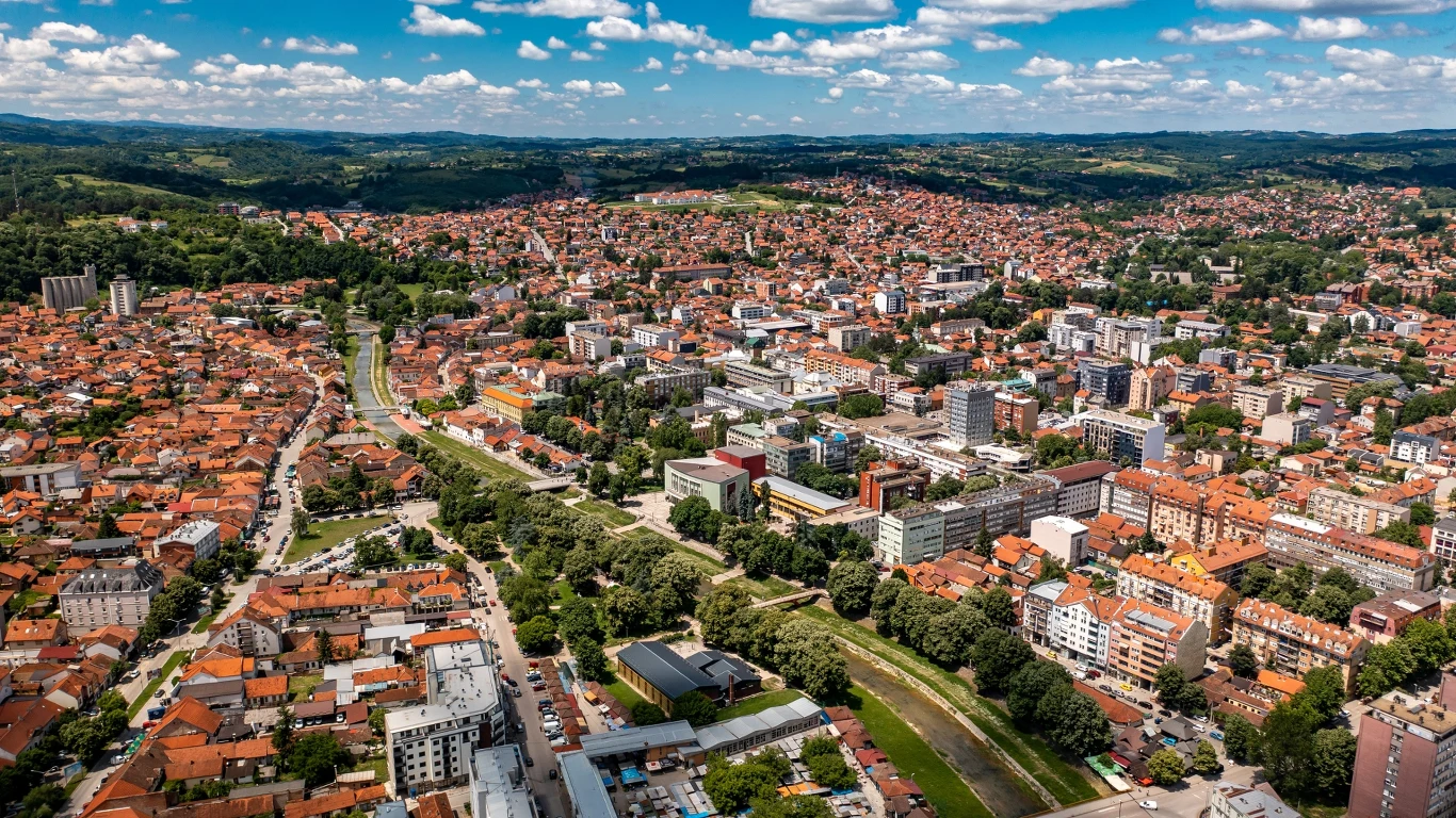 Valjevo Rozległa panorama miasta z ceglanymi dachami, zielonymi drzewami i wijącą się przez środek rzeką, otoczona niską, rozproszoną zabudową mieszkaniową oraz kilkoma nowoczesnymi budynkami wyróżniającymi się spośród tradycyjnej architektury.