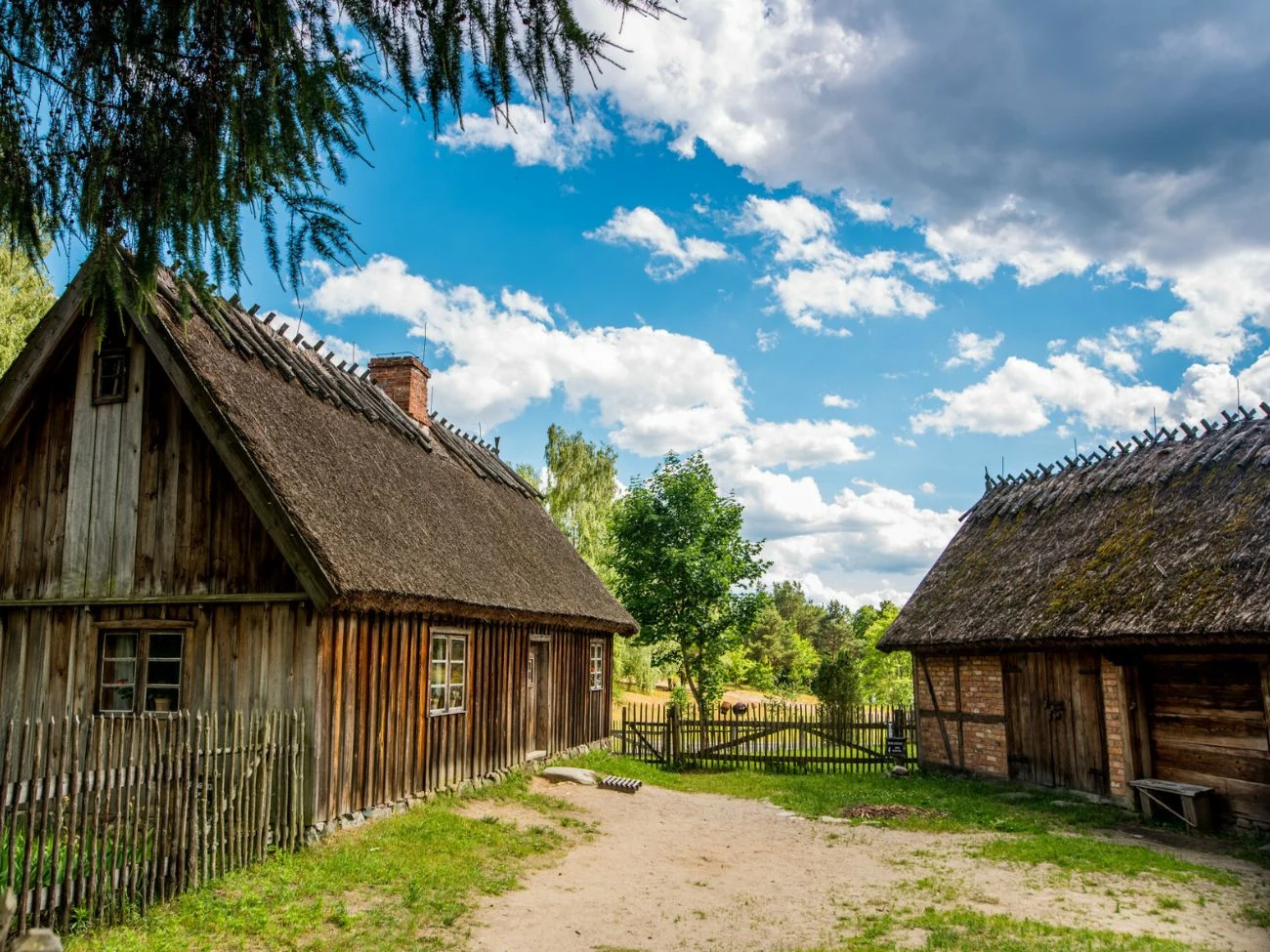 Kaszubski Park Etnograficzny - Wdzydze Kiszewskie Drewniane chaty kryte strzechą w Kaszubskim Parku Etnograficznym we Wdzydzach, otoczone płotem, z drzewami w tle.