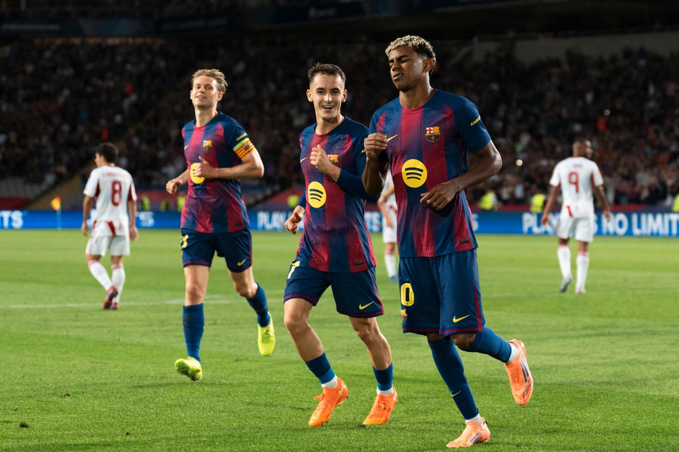 Trzech piłkarzy FC Barcelony w granatowo-bordowych strojach biegnie po murawie stadionu, wyrażając radość po zdobyciu bramki podczas meczu, w tle widoczni zawodnicy drużyny przeciwnej oraz publiczność na trybunach.