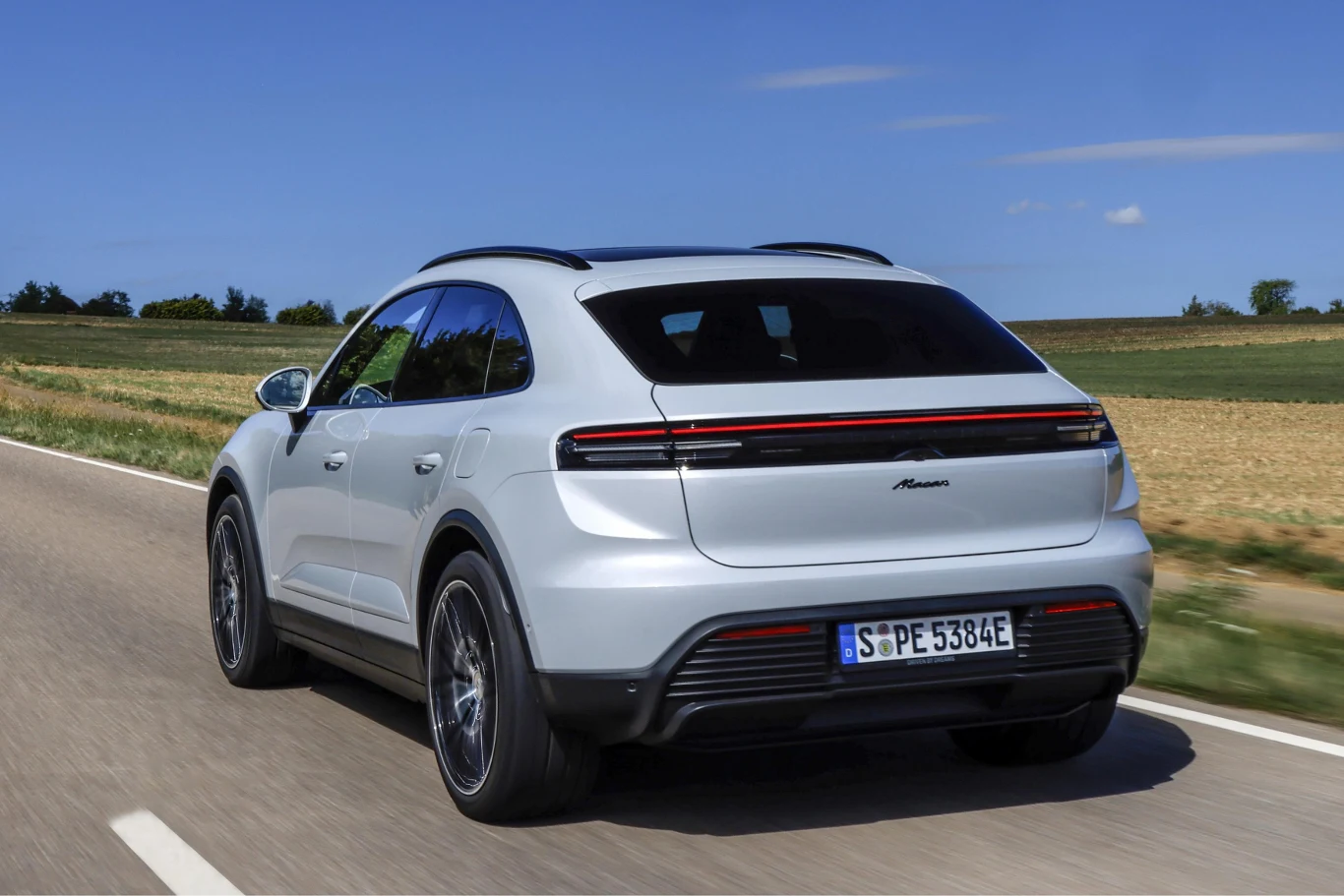 Srebrny SUV o nowoczesnym designie z panoramicznym dachem i charakterystyczną, ciągłą listwą świetlną z tyłu jadący drogą pośród pól przy słonecznej pogodzie.