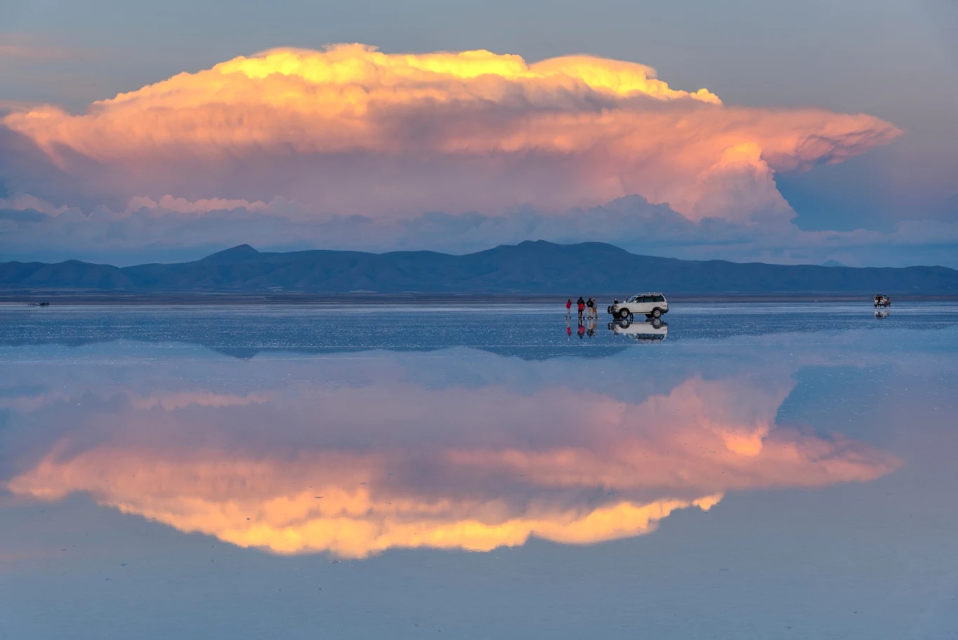 Widoki na Salar de Uyuni w porze deszczowej. Ogromna, podświetlona chmurami formacja odbijająca się w wodzie solniska, pośród której stoi samochód terenowy i kilka postaci na tle gór.