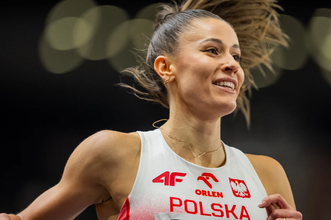 Uśmiechnięta polska lekkoatletka w stroju z napisem ‘Polska’ i charakterystycznymi logotypami, podczas biegu na zawodach sportowych.