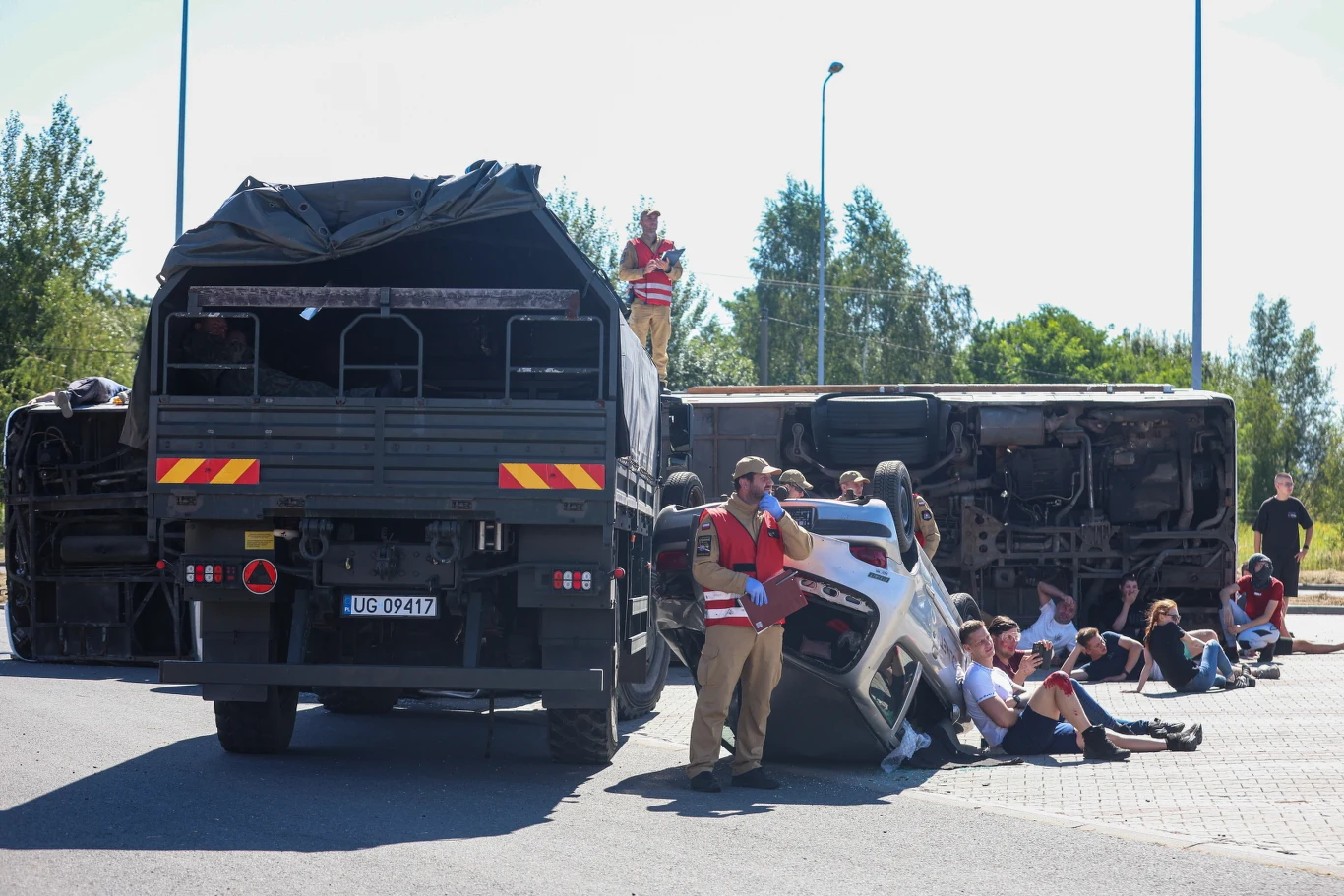 Taran na autostradzie A1 Przewrócony autobus leżący na boku, wojskowy samochód ciężarowy i uszkodzony biały samochód osobowy, wokół których zgromadzona jest grupa ludzi, część siedzi lub klęczy na ziemi, a jedna osoba w kamizelce odblaskowej udziela pomocy.