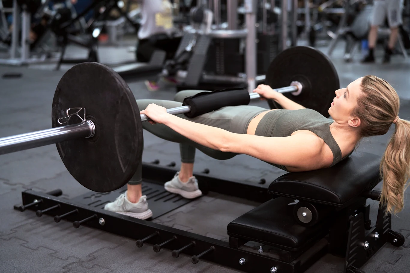 Kobieta w sportowym stroju wykonuje hip thrust z użyciem ciężkiej sztangi na specjalnej ławce na siłowni.
