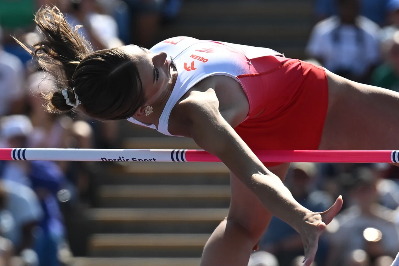 Maja Słodzińska Zawodniczka w czerwono-białym stroju sportowym pokonuje poprzeczkę podczas skoku wzwyż, z wyraźnym wysiłkiem i skupieniem na twarzy.