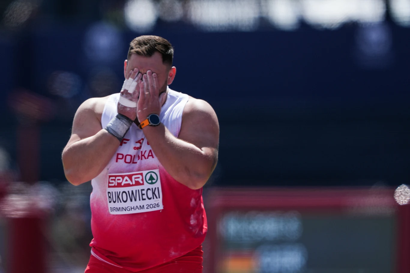 Konrad Bukowiecki Lekkoatleta w stroju reprezentacji Polski o umięśnionej sylwetce zakrywa twarz dłońmi, na piersi widoczny numer startowy z nazwiskiem oraz informacją o zawodach w Birmingham 2026.