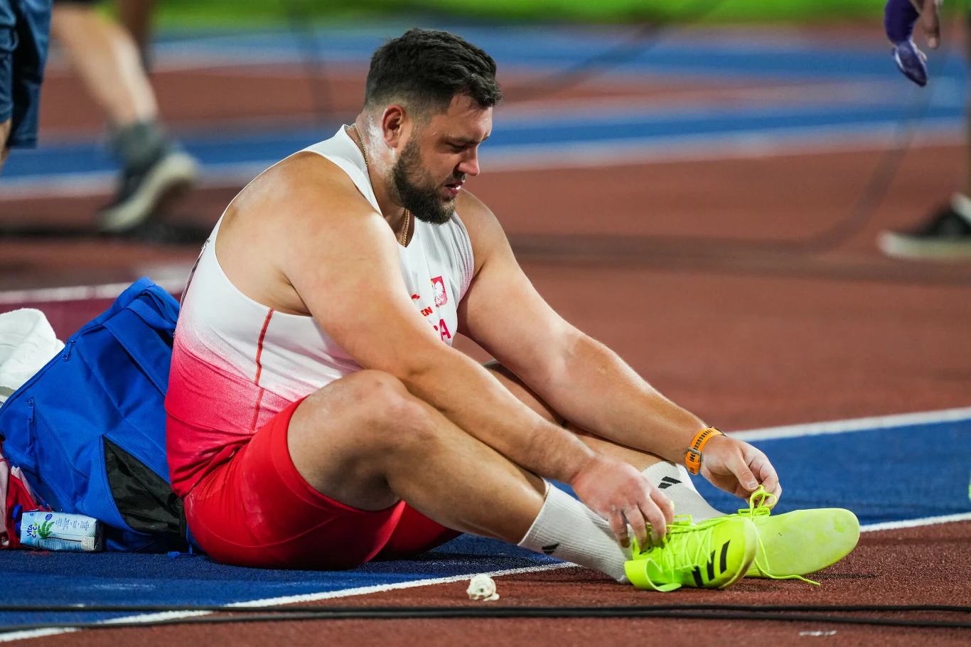 Konrad Bukowiecki Zawodnik w sportowym stroju siedzi na bieżni i wiąże jasnożółte buty, obok plecak i butelka z wodą.