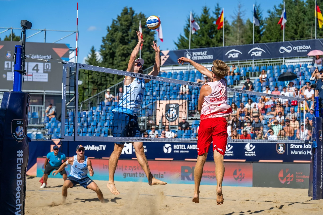 W ataku Aleksander Czachorowski Dwóch siatkarzy plażowych walczących o piłkę nad siatką podczas meczu, jeden ubrany w niebieski strój blokuje atak zawodnika w czerwonym stroju, na tle licznej widowni na trybunach.