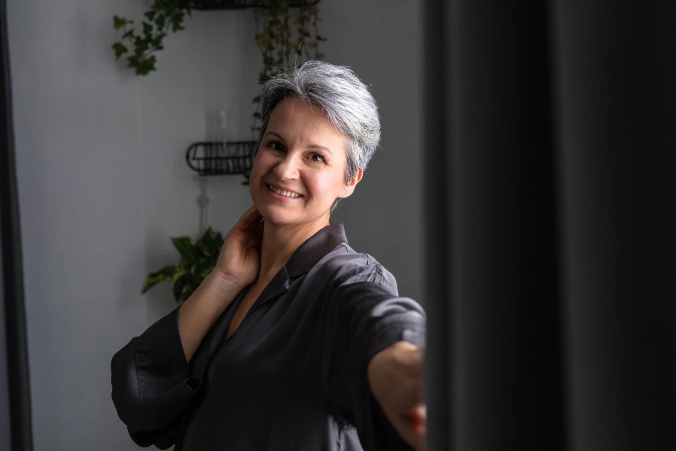 Co sprawia, że twarz wygląda młodo? Nie chodzi tylko o brak zmarszczek Uśmiechnięta kobieta z krótkimi, siwymi włosami w ciemnej koszuli stoi przy oknie, trzymając się za kark i spoglądając w obiektyw na tle roślin doniczkowych.