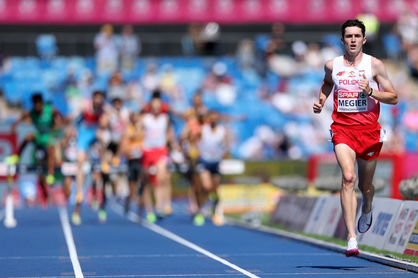 Polski lekkoatleta w czerwono-białym stroju wyraźnie prowadzi w biegu na stadionie, podczas gdy pozostali zawodnicy znajdują się daleko za nim na niebieskiej bieżni.