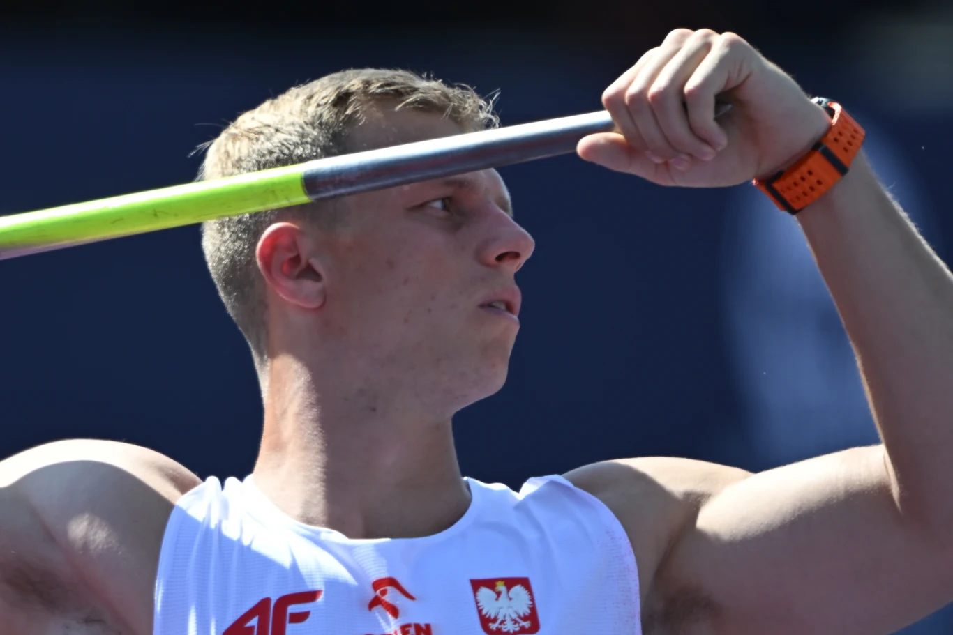 Dawid Wegner Lekkoatleta w białej koszulce z orłem i polskim godłem przygotowuje się do rzutu oszczepem, skupiony, z uniesionym ramieniem i oszczepem opartym o bark.