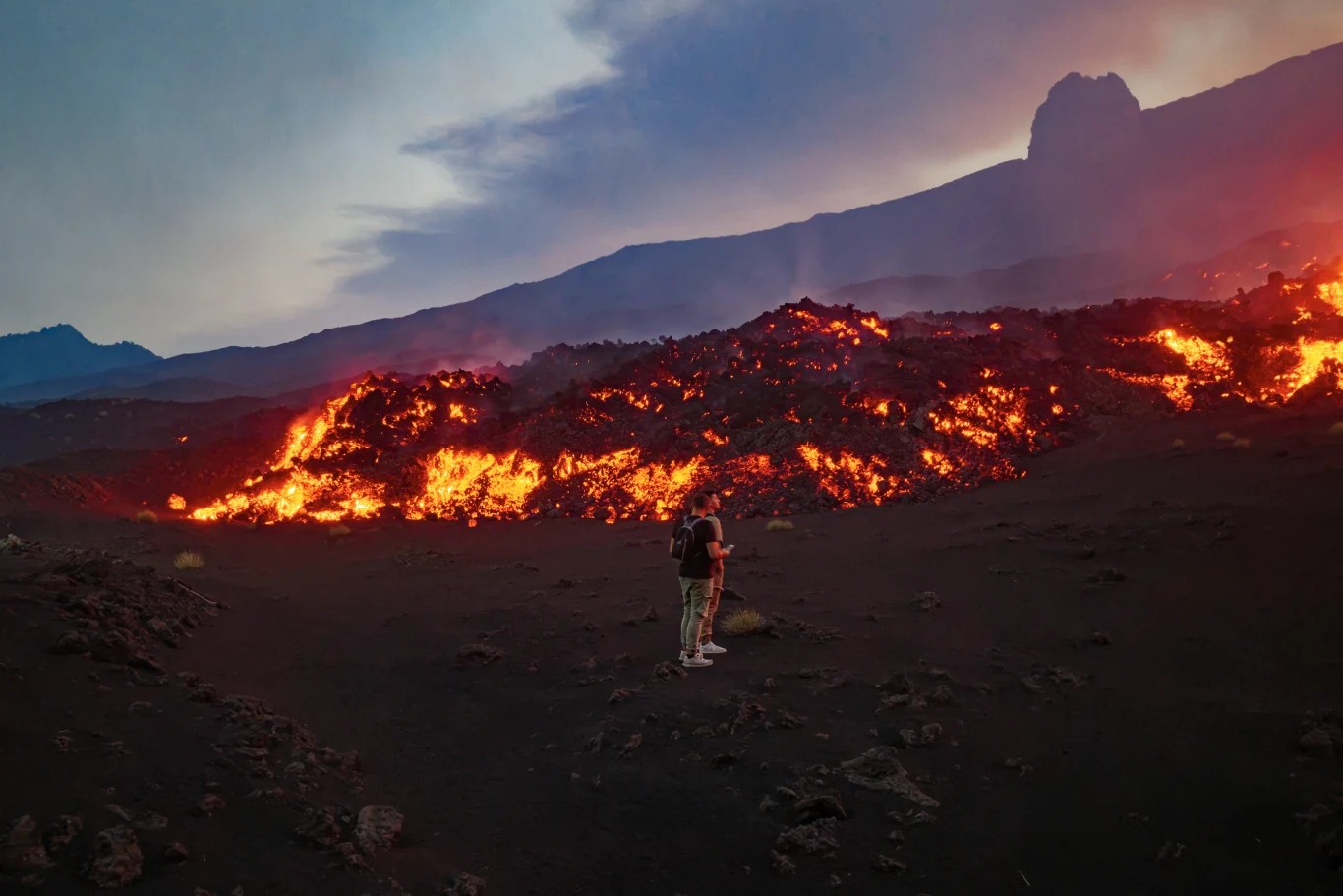 Setki turystów chcą podziwiać erupcję wulkanu Etna Turysta stoi na ciemnych skałach obok potoku czerwonej lawy wypływającej z wulkanu Etna.