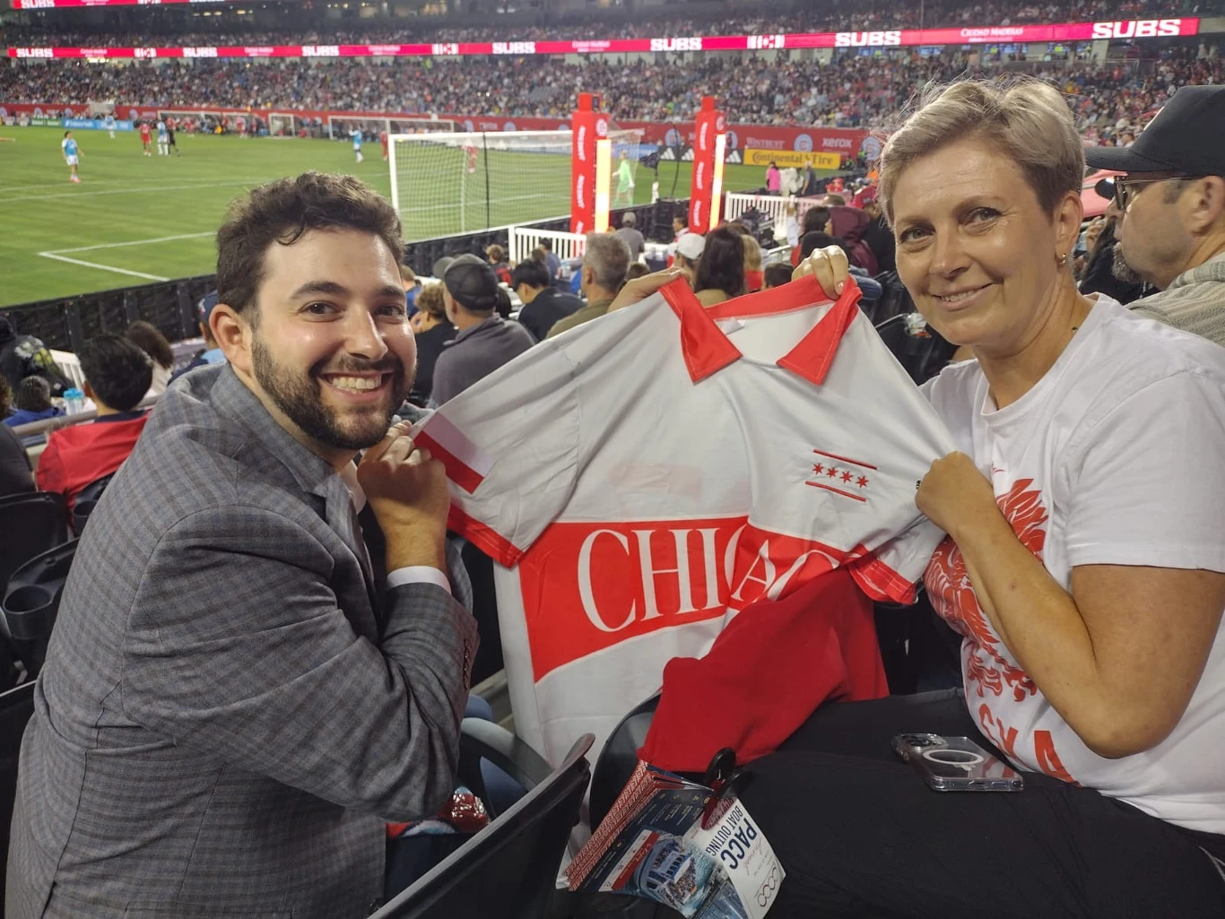 Monika Wyrwas podczas meczu Chicago Fire - Charlotte Dwoje uśmiechniętych kibiców siedzi na trybunach stadionu piłkarskiego i prezentuje biało-czerwoną koszulkę z napisem „CHICAGO” oraz czterema gwiazdkami, w tle trwa mecz piłki nożnej przy pełnych trybunach.