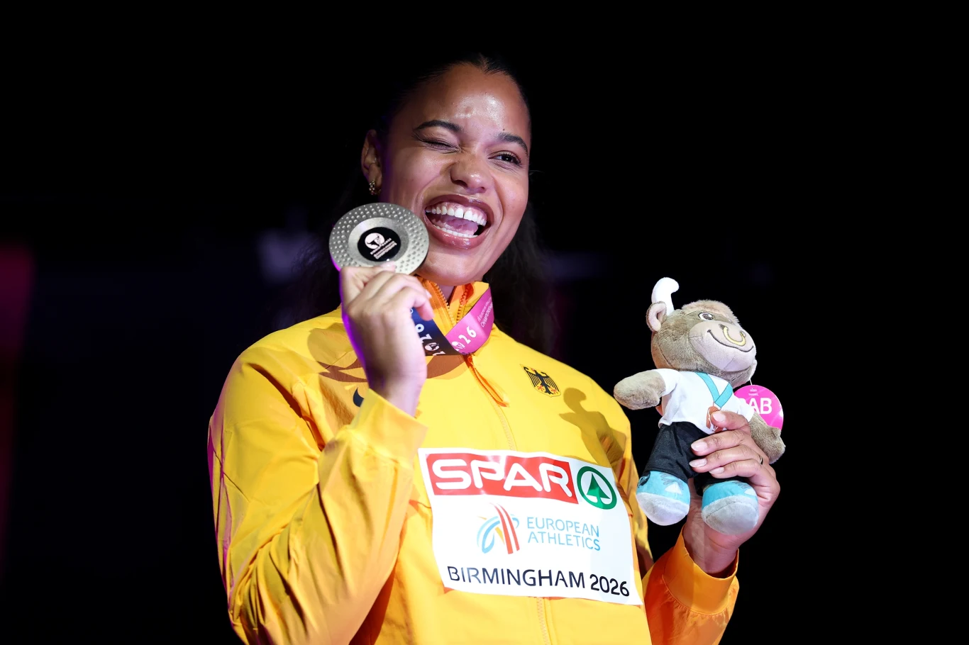 Yemisi Mabry Uśmiechnięta sportsmenka w żółtym dresie prezentuje srebrny medal oraz maskotkę na tle ciemnego tła, trzymając medal na szyi i unosząc go w geście zwycięstwa.