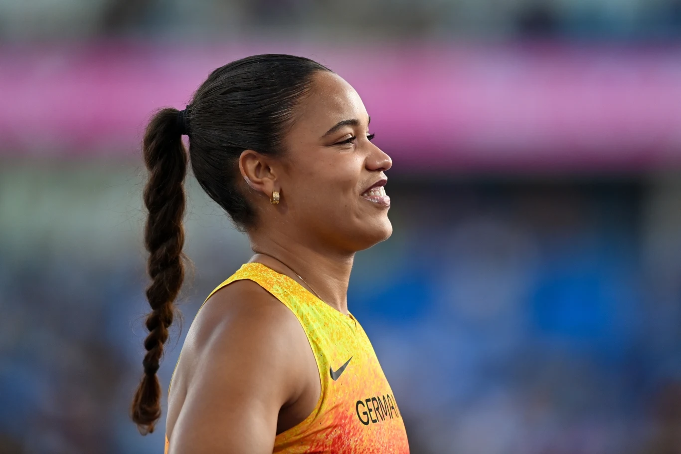 Yemisi Mabry Uśmiechnięta lekkoatletka w żółto-pomarańczowym stroju z napisem Germany i długim warkoczem, na tle rozmytej areny sportowej.