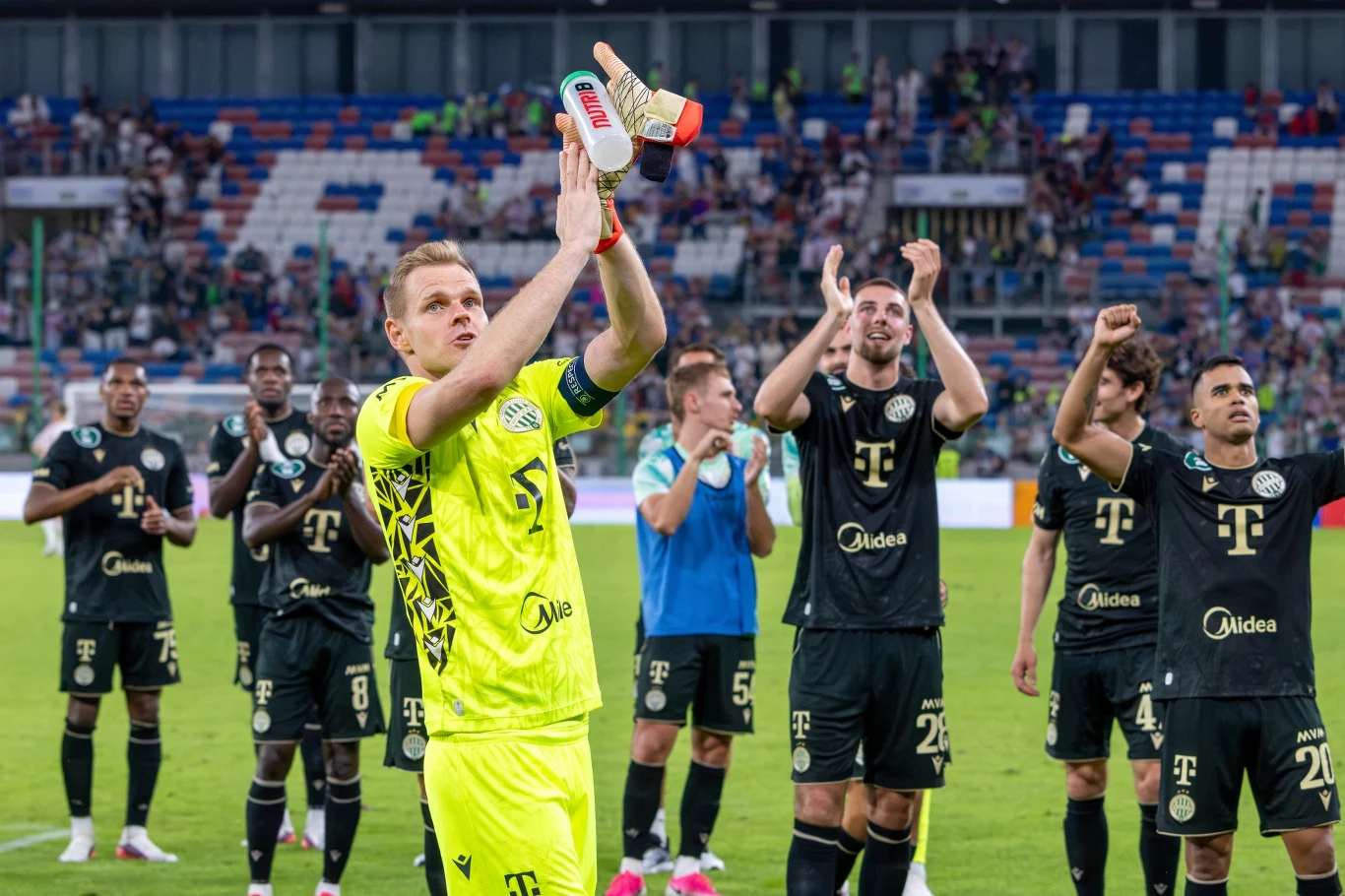 Grupa piłkarzy klubu w ciemnych strojach dziękuje kibicom na stadionie, bramkarz w jasnożółtym komplecie klaska z butem w dłoni.