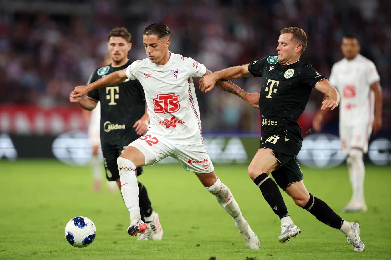 Kacper Urbański (z lewej) otworzył wynik spotkania Górnik - Ferencvaros (1:1) Dwóch piłkarzy walczących o piłkę na boisku podczas meczu, jeden w białym stroju prowadzi futbolówkę, drugi w czarnym próbuje odebrać ją rywalowi.