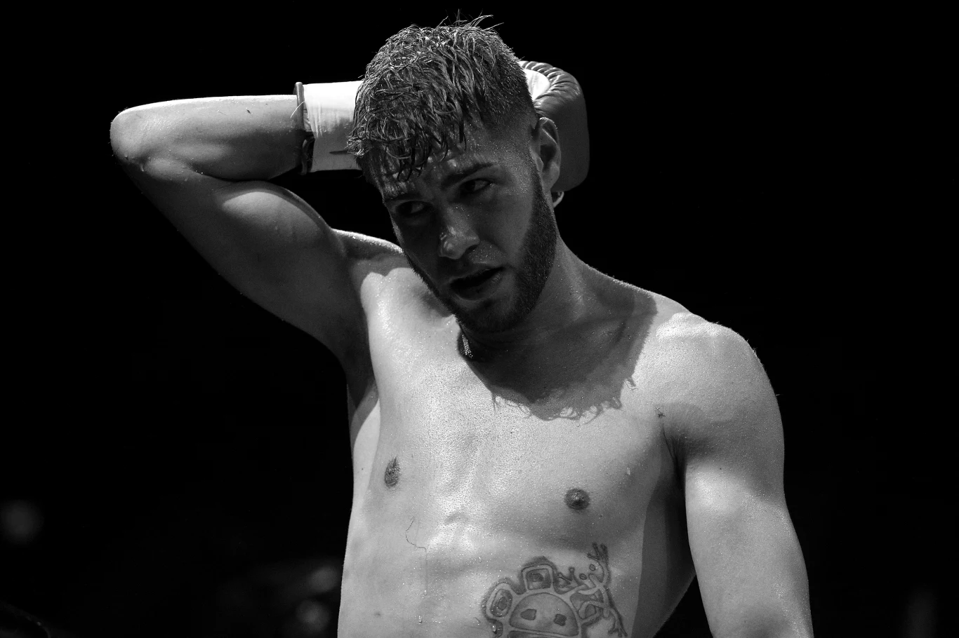 Prichard Colon Zmęczony, umięśniony mężczyzna z tatuażem na klatce piersiowej odgarnia mokre włosy ręką, stojąc na ciemnym tle.