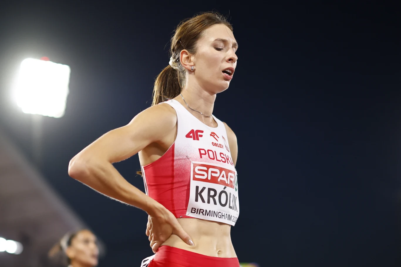 Kinga Krółik Lekkoatletka w stroju reprezentacji Polski z numerem startowym, lekko pochylona do przodu, z ręką opartą na biodrze, w wyrazie zmęczenia po zawodach sportowych na stadionie oświetlonym reflektorami.