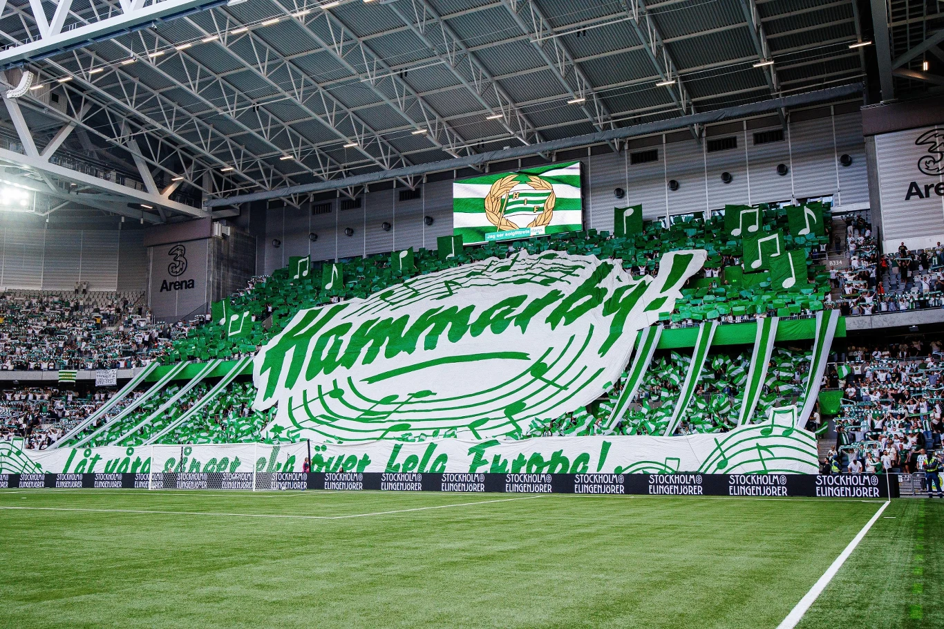 Stadion Hammarby Stadion Hammarby, kartoniada w zielono-białych barwach z nutami i nazwą klubu, kibice na trybunach.