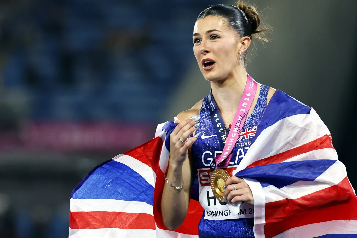Lekkoatletka z Wielkiej Brytanii w stroju sportowym owinięta brytyjską flagą, trzymająca złoty medal i różowy szalik z napisem będąca na stadionie po zakończonych zawodach.