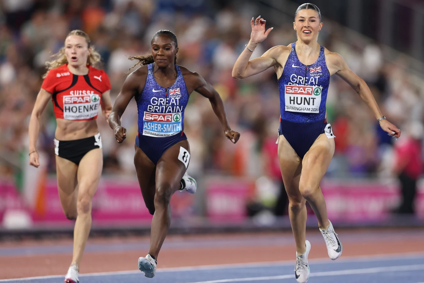 Trzy zawodniczki biegnące na finiszu biegu sprinterskiego na stadionie lekkoatletycznym, z przewagą jednej z nich w niebieskim stroju reprezentującym Wielką Brytanię.