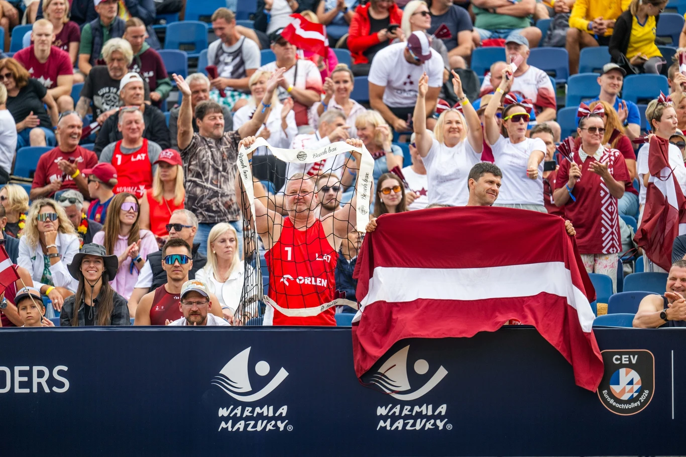 Kibice na trybunach ubrani w barwy narodowe, duża flaga Łotwy zawieszona na barierce, atmosfera sportowego wydarzenia z dopingującym tłumem.