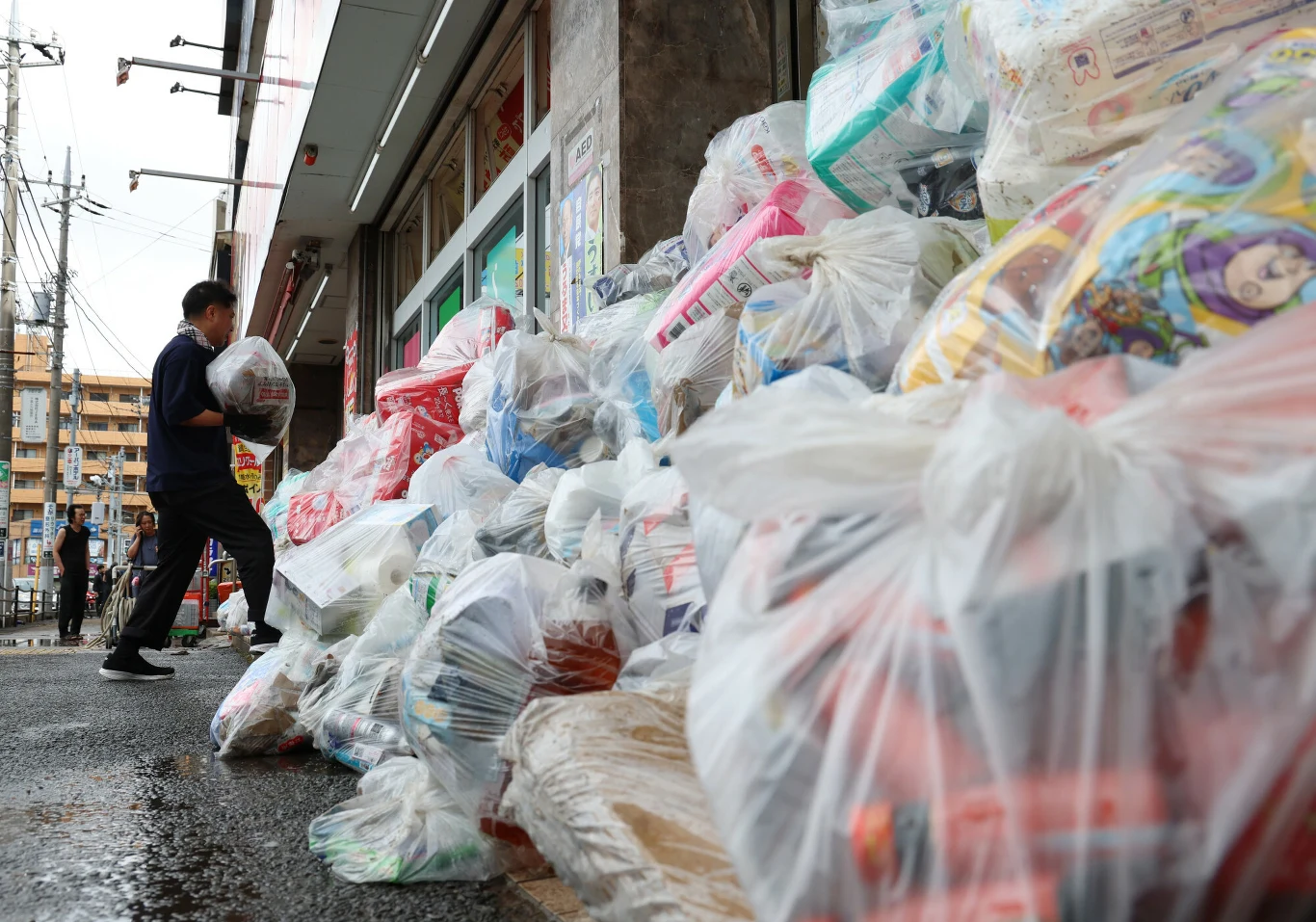 Japonia sprząta po wielkich ulewach i powodzi Stosy worków z odpadami ułożone przed wejściem do sklepu spożywczego, mężczyzna przenoszący kolejny worek na mokrym chodniku.