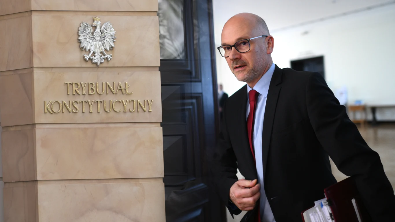 Mężczyzna w okularach i garniturze przechodzi obok marmurowej ściany z napisem 'Trybunał Konstytucyjny' oraz godłem Polski, trzymając pod pachą dokumenty.