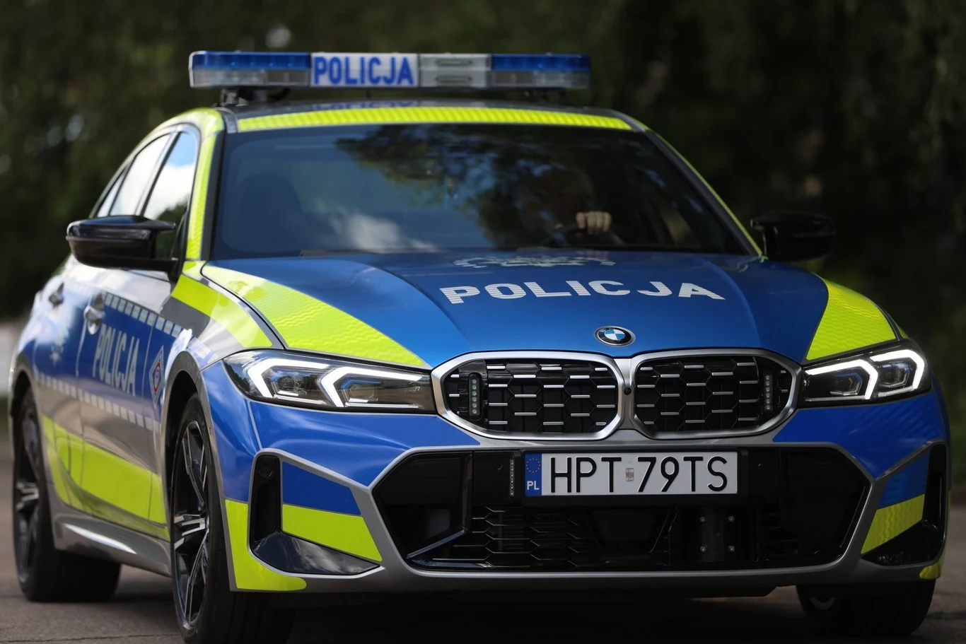BMW M340i xDrive wyposażone jest w sześciocylindrowy, trzylitrowy silnik benzynowy generujący 381 KM. Wspomaga go układ miękkiej hybrydy o mocy 12 KM. Razem auto dysponuje mocą 392 KM. Radiowóz marki BMW w biało-niebieskich barwach z widocznym napisem "Policja" i charakterystycznym oznakowaniem, wyposażony w światła ostrzegawcze na dachu, widziany od przodu na tle zieleni.