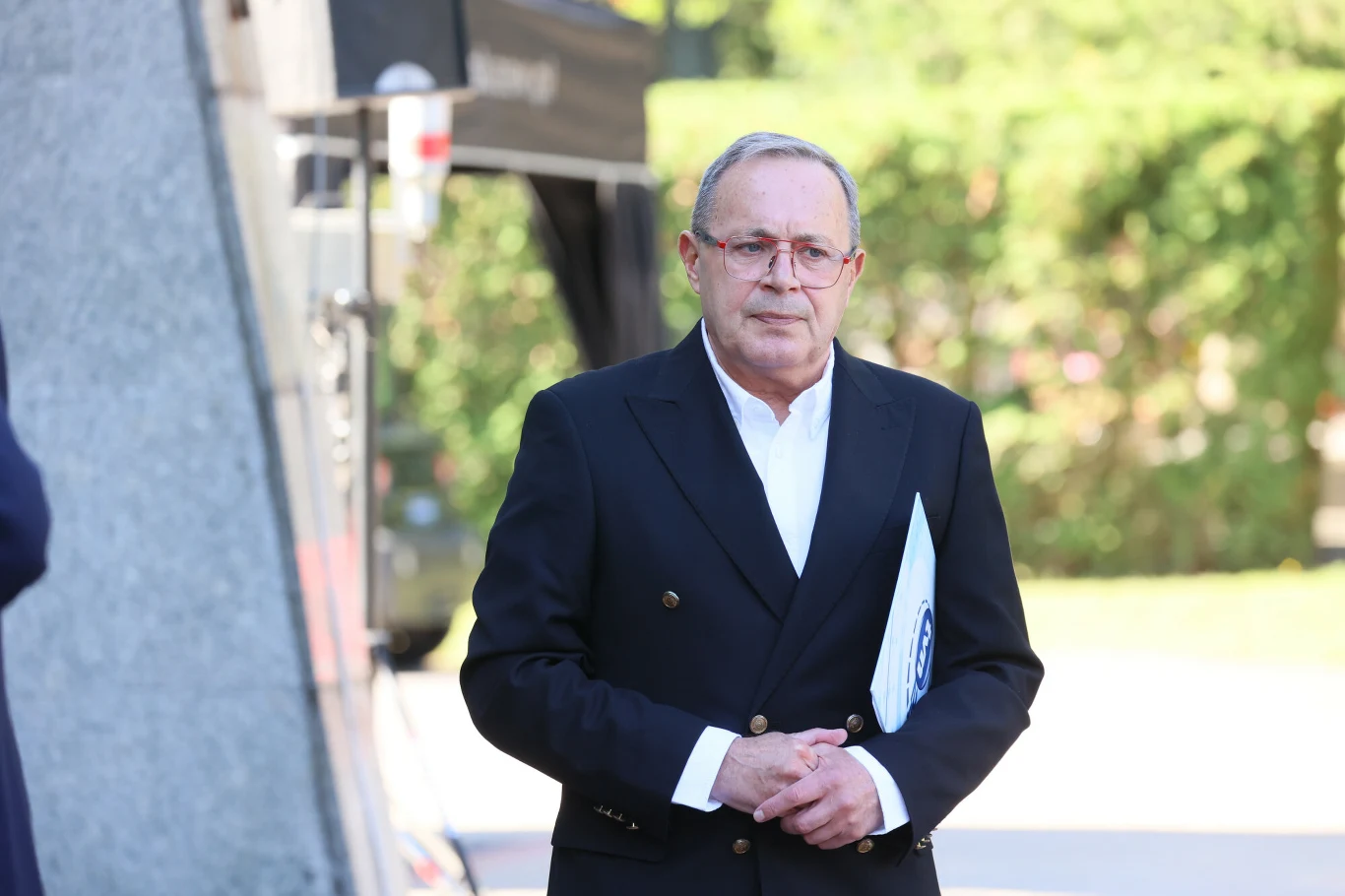 Mężczyzna w eleganckim, ciemnym garniturze ze złotymi guzikami i białą koszulą trzyma dokument, stojąc na świeżym powietrzu, z zielenią w tle.