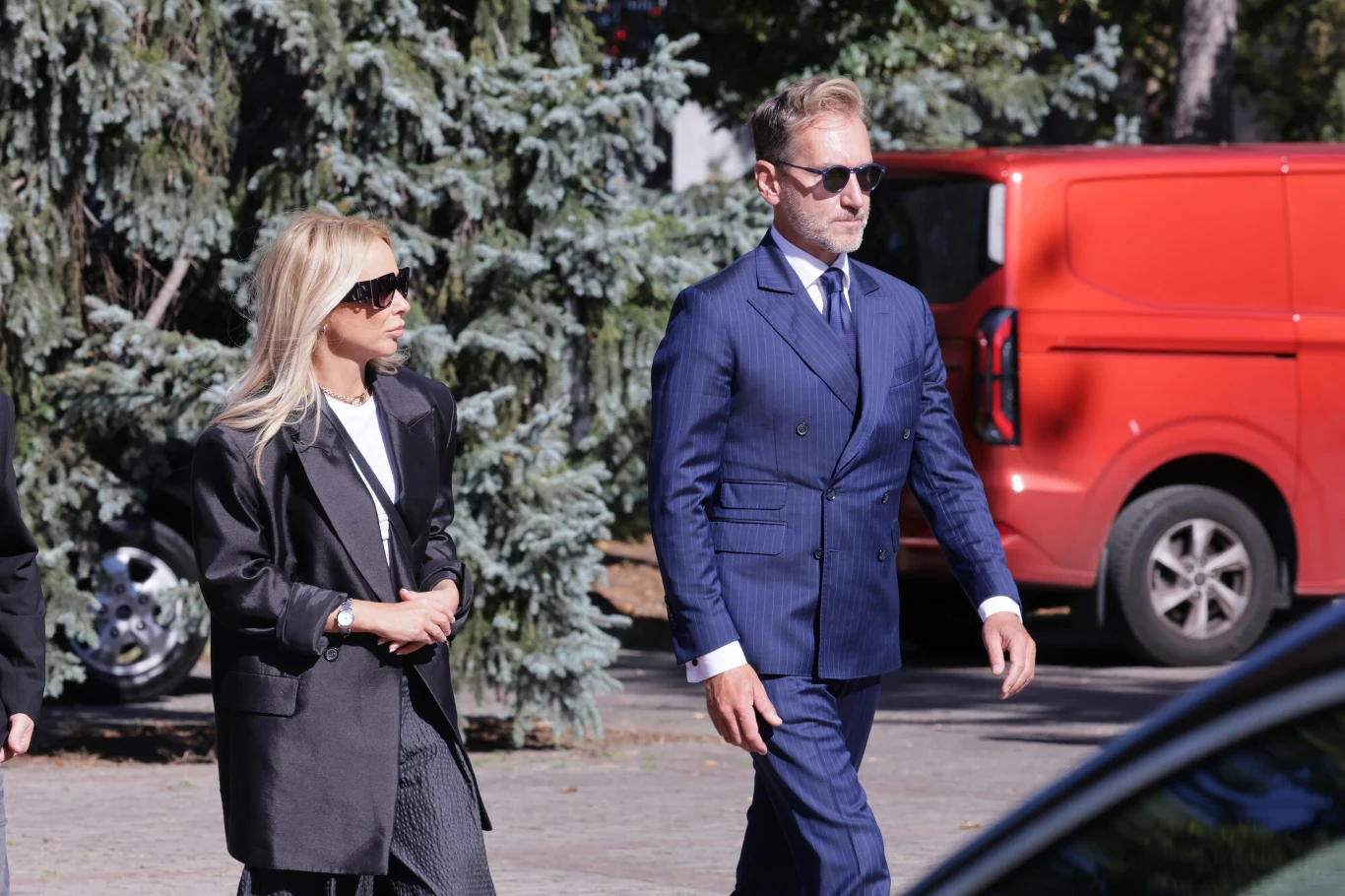 Dwójka elegancko ubranych osób idzie razem chodnikiem; kobieta w ciemnych okularach i oversize'owej marynarce oraz mężczyzna w granatowym garniturze i okularach przeciwsłonecznych, w tle czerwona furgonetka i zielone drzewa.