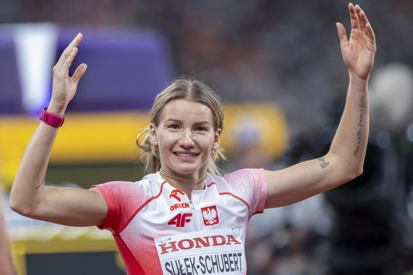 Adrianna Sułek-Schubert Lekkoatletka w biało-czerwonym stroju z napisem "Sulek-Schubert" i emblematami sponsorów podnosi ręce w geście triumfu, uśmiechając się szeroko.