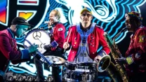 Czterech muzyków w kolorowych mundurach gra na instrumentach dętych i perkusyjnych na tle dynamicznej scenografii świetlnej.