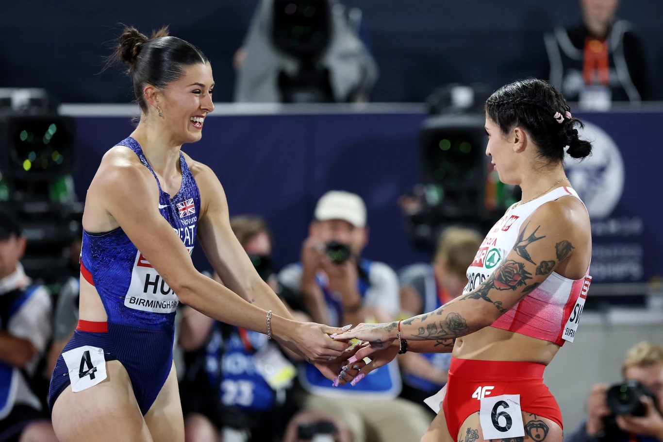Dwie lekkoatletki w sportowych strojach ściskają sobie dłonie po biegu, wymieniając uśmiechy na tle fotografujących ich osób.