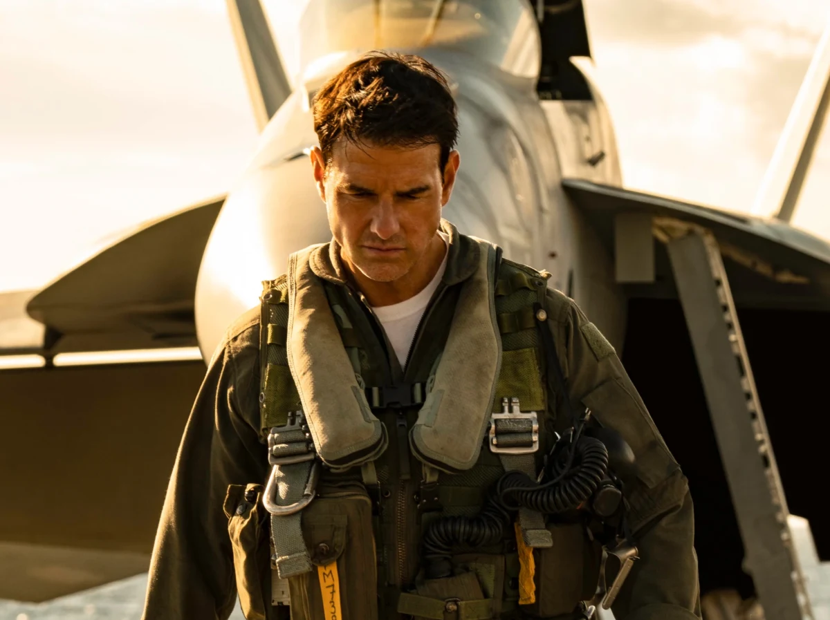 Tom Cruise w filmie Top Gun: Maverick Tom Cruise w kombinezonie lotniczym i kamizelce pilota stojący przed wojskowym myśliwcem na zewnątrz.