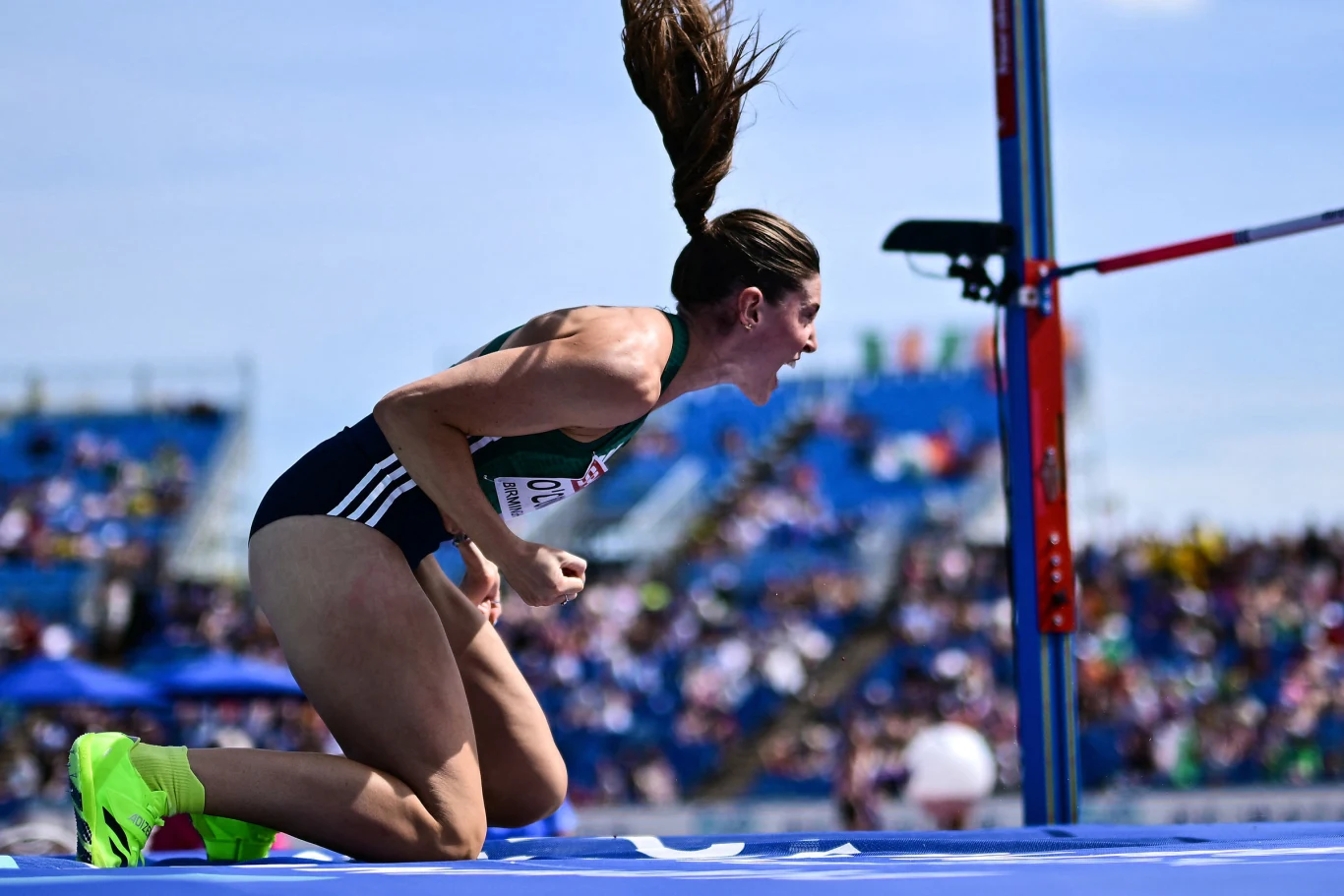 Kate O'Connor Zawodniczka w sportowym stroju i neonowych butach klęczy na bieżni obok poprzeczki do skoku wzwyż, zaciskając pięści i wyrażając silne emocje po wykonaniu próby, w tle trybuny z widzami.