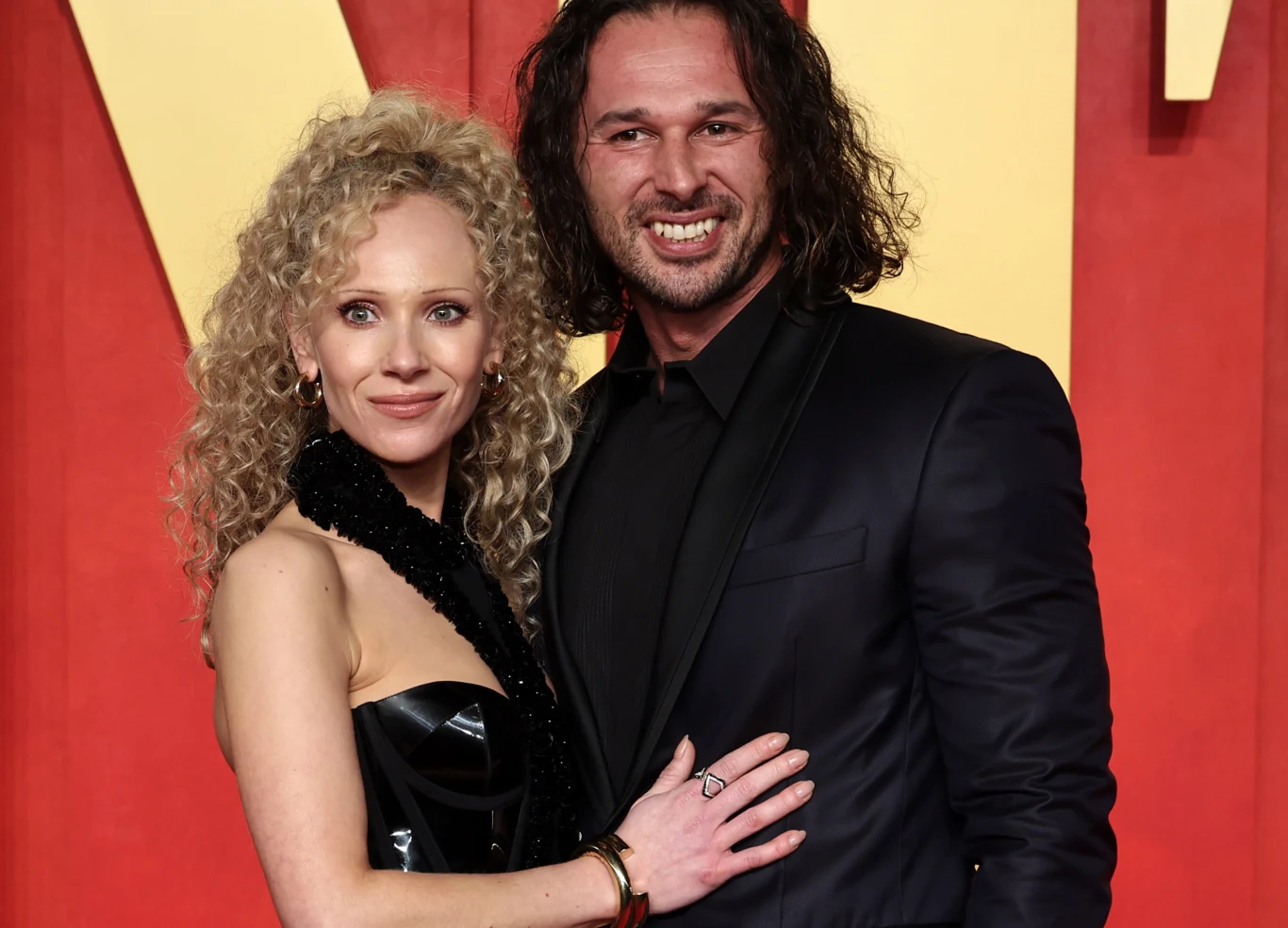 Juno Temple, Michał Szymański Kobieta z blond, kręconymi włosami w czarnej sukni i mężczyzna z długimi, ciemnymi włosami ubrany w czarny garnitur pozują razem, obejmując się i uśmiechając do kamery na tle czerwonej i żółtej dekoracji.