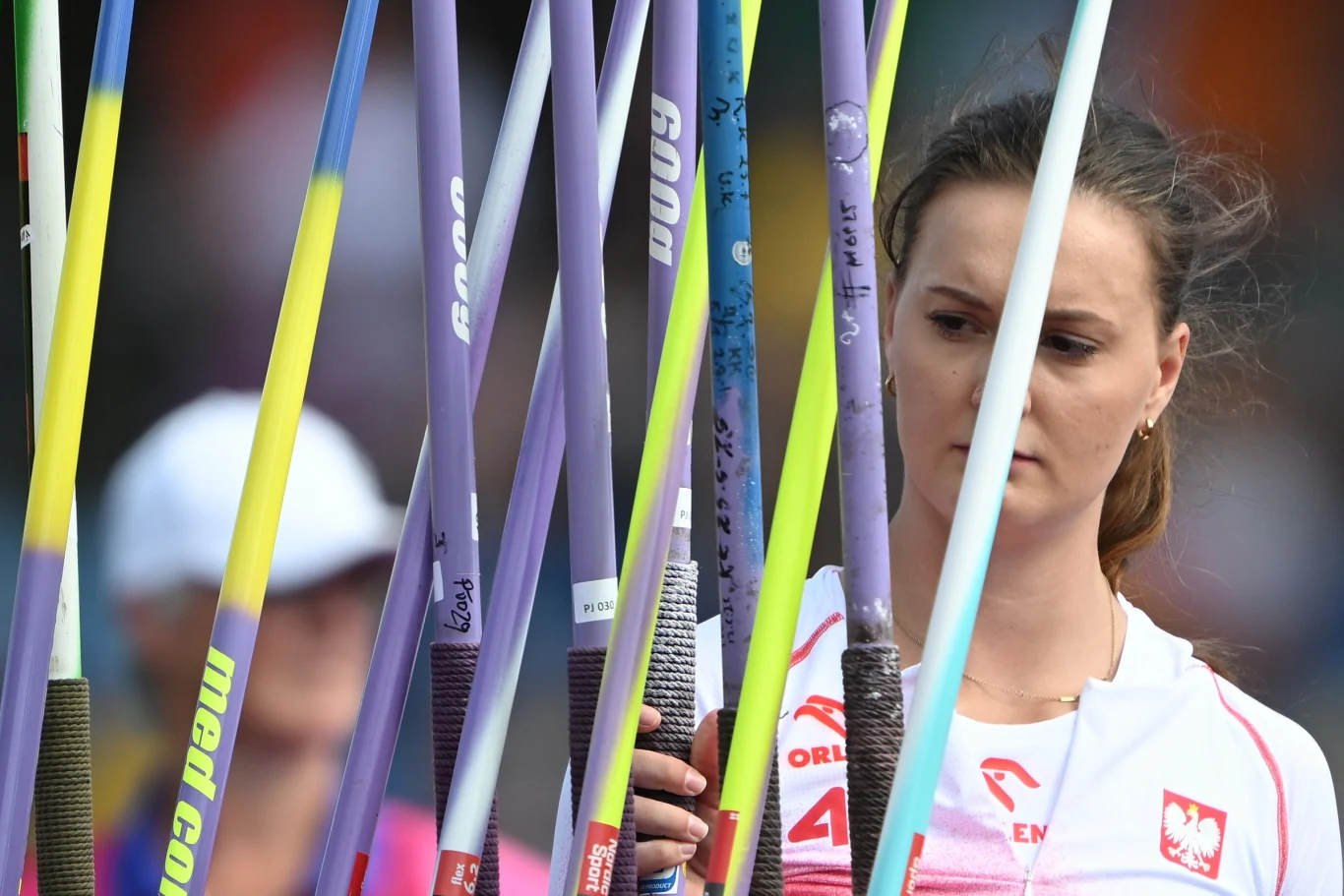 Zawodniczka w stroju reprezentacji Polski wybiera oszczep spośród wielu kolorowych sprzętów sportowych ustawionych pionowo przed sobą.