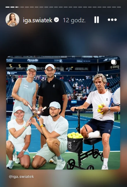 Iga Świętek z teamem i doklejonym Francisco Roigiem Grupa pięciu osób na korcie tenisowym, wszyscy uśmiechnięci, w sportowych ubraniach, jedna osoba siedzi na urządzeniu z piłkami tenisowymi, pozostałe trzymają wspólnie małą statuetkę.
